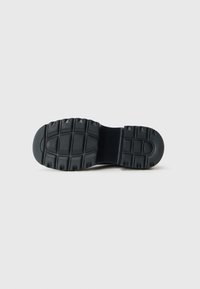 Black shoe sole with deep tread pattern featuring horizontal and vertical grooves for traction, against a plain light gray background.