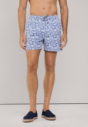 Costumi da bagno blu e bianchi con un motivo a foglie, cintura elastica e cordoncino. Il modello indossa scarpe slip-on blu navy.
