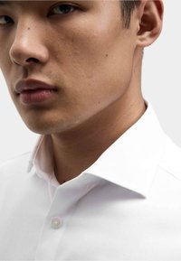 Nahaufnahme des Kieferlinien und der Lippen eines Mannes, der ein weißes Hemd mit Kragen trägt, vor einem einfarbigen hellen Hintergrund.