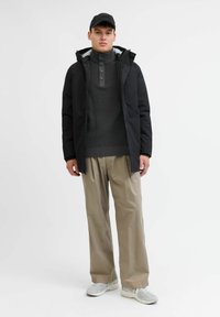Chaqueta negra con capucha, suéter gris de punto con cuello alto, pantalones beige de pierna ancha y zapatillas ligeras. La modelo está de pie contra un fondo blanco sencillo.