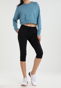 Felpa blu tagliata con logo testurizzato, abbinata a leggings neri a tre quarti e scarpe da ginnastica bianche, evidenziando un fit rilassato e comfort.