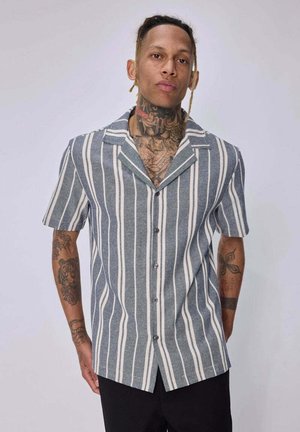 Homme avec des tatouages au cou et aux bras porte une chemise à manches courtes boutonnée avec des rayures verticales blanches et grises sur fond uni.