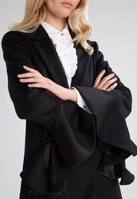 Sort skræddersyet jakke med brede, flagrende ærmer; luksuriøst stof; hvid ruflet bluse under; gyldne knapper på kraven.