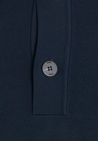 Tissu en maille bleu marine présentant un motif texturé, avec un détail de bouton argenté portant l'inscription "HUGO" et une étiquette en tissu rectangulaire à côté du bouton.