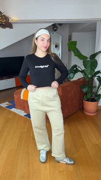 Tânără cu păr lung, purtând un top crop negru cu mâneci lungi, pantaloni bej, pantofi argintii și o bentiță albă, stând într-un living.