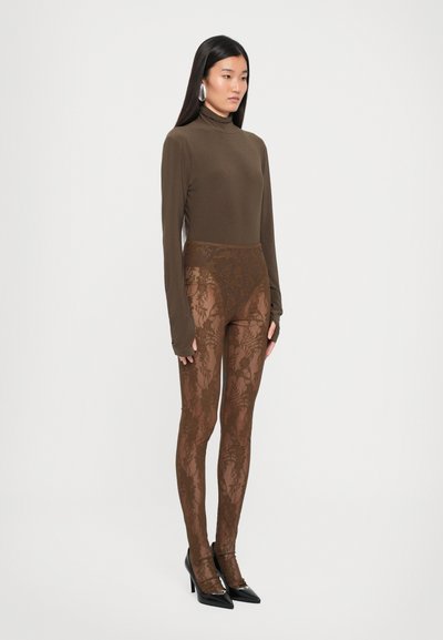 Brun langermet topp kombinert med semi-transparent brune blomstermønstrede leggings i blonder. Modellen har på seg spisse svarte hæler, noe som fremhever outfitens strukturerte elementer.