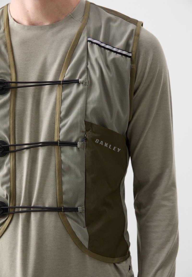 Homme portant un gilet en maille beige avec des fermetures à cordon élastique noir et une poche latérale olive étiquetée "Oakley" sur une chemise à manches longues.