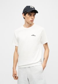 Νέος άνδρας που φοράει λευκό μπλουζάκι με το κείμενο "OBEY WORLDWIDE", ανοιχτό γκρι σορτς και μαύρο καπέλο μπέιζμπολ με ρίγες, κοιτάζοντας πλάγια.