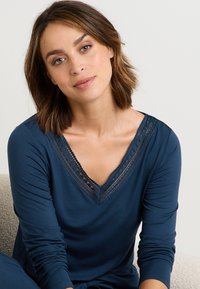 Haut à manches longues bleu marine avec un col en V orné de dentelle noire, en tissu lisse et avec des manches ajustées. Design minimaliste avec des détails structurés au niveau du col.