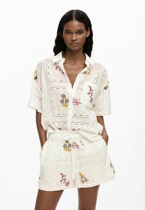 Mujer con camisa blanca de manga corta abotonada y shorts a juego con bordados coloridos de flores y pájaros, con las manos en los bolsillos.