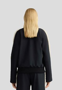 Giacca bomber nera con pannelli laterali in crema morbida e testurizzata e polsini a costine. Materiale liscio con una vestibilità rilassata, adatta per essere indossata a strati.