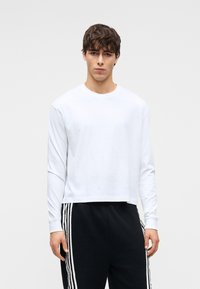 Λευκό μακρυμάνικο T-shirt από βαμβακερό υλικό, με στρογγυλή λαιμόκοψη και άνετη εφαρμογή. Συνδυασμένο με μαύρη φόρμα με άσπρες ρίγες.