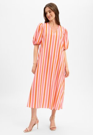 Femme portant une robe midi à rayures verticales roses et orange avec des manches bouffantes, assortie à des sandales à talons hauts couleur nude, debout sur fond blanc.