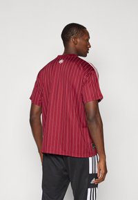 Mand står med ryggen til iført en maroon, hvidstribet kortærmet skjorte og sorte Adidas træningsbukser med hvide striber ned langs siderne.