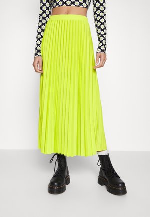 Jupe maxi plissée jaune néon portée avec un crop top noir à fleurs à manches longues et des bottes de combat noires à lacets avec des chaussettes blanches.