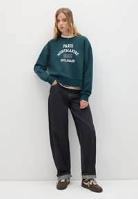 Joven mujer con cabello rubio, vistiendo un sudadera verde de París Montmartre, jeans anchos negros y zapatillas marrones, de pie contra una pared blanca.