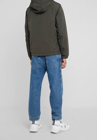 Olive green hooded jacket, lightweight material, paired with baggy light blue jeans, and white sneakers with gray accents and logo detail.