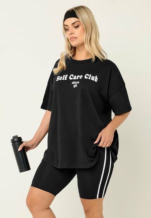 Frau in schwarzem Sportoutfit und Stirnband hält eine schwarze Wasserflasche, trägt ein lockeres T-Shirt mit dem Aufdruck „Self Care Club seit 96“.