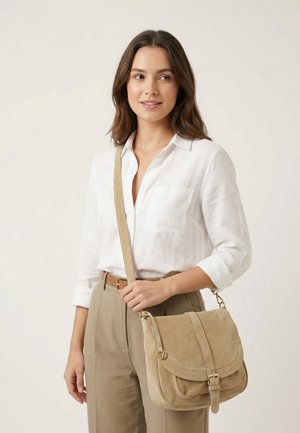 Femme portant une chemise blanche boutonnée et un pantalon beige, avec un sac bandoulière en daim couleur noisette orné d'une boucle.
