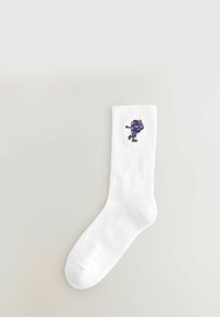 Hvid crew-sok med ribbet manchet, der har en lille broderet lilla blomstfigur, der ifører sneakers på siden.