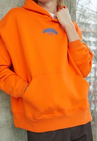 Sudadera con capucha naranja y bolsillo frontal, con el texto azul "WRSTBHVR STUDIO" bordado en el pecho, fabricada en un tejido suave.
