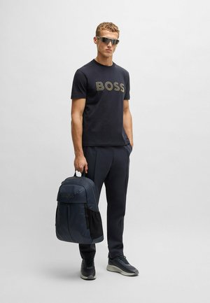 Navy backpack with logo, worn with a black t-shirt featuring a large gold "BOSS" print, paired with dark athletic pants and shoes.