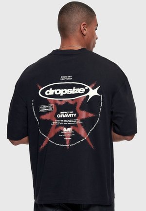 Bărbat purtând un tricou negru cu text grafic alb și roșu despre gravitație și marca dropsize pe spate.