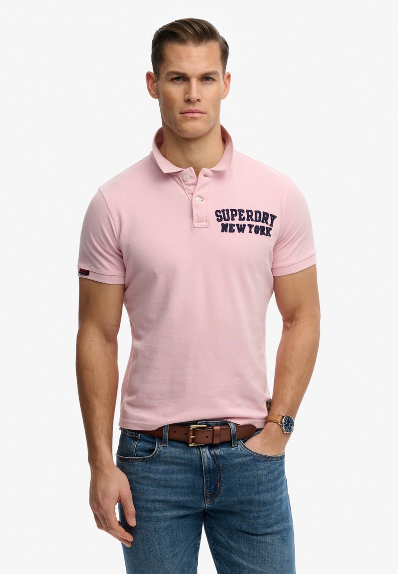 Mann trägt ein hellrosa Poloshirt mit dem Text "Superdry New York", blaue Jeans, braunen Gürtel und eine Armbanduhr, steht mit einer Hand in der Tasche.