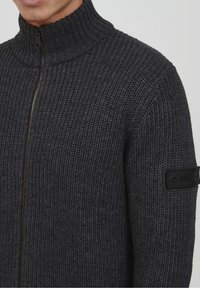 Pull en tricot gris foncé avec un col haut et une fermeture éclair sur toute la longueur. Présente un motif texturé et un patch noir de marque sur la manche.