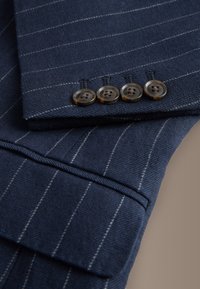 Navy blue pinstripe suit sleeve with four dark brown buttons and a flap pocket on a beige background.