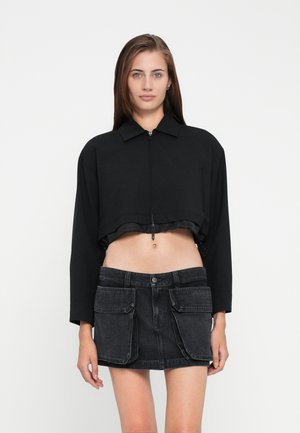 Zwarte cropped jas met een kraag en ritssluiting, gecombineerd met een donkere denim rok met opgestikte zakken.