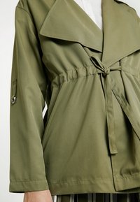 Oversized jakke i olivengrøn med brede revers, justerbart bælte i taljen og lange ærmer med en løkke-detalje på ærmet. Glat tekstur.