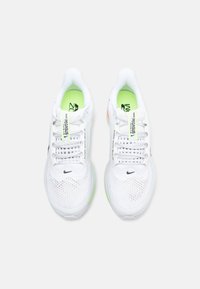 Λευκά αθλητικά παπούτσια με ύφασμα από δίχτυ, πράσινες λεπτομέρειες, γκρι και πορτοκαλί στοιχεία, και το μαύρο λογότυπο Nike κοντά στις κορδόνια.