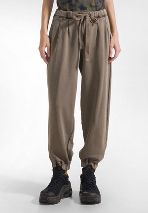 Persona que lleva pantalones jogger marrones con cordón y puños elásticos, y zapatos deportivos negros, de pie frente a un fondo liso.