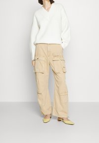 Pull en tricot blanc à côtes avec un décolleté en V, associé à un pantalon cargo beige doté de multiples poches et d'une coupe décontractée. Chaussures à talons bloc jaunes.