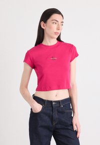 Mladá žena sa pózuje, oblečená v priliehavom ružovom crop topu s logom, skombinovanom s tmavými džínsami. Stojí proti neutrálnej pozadí.