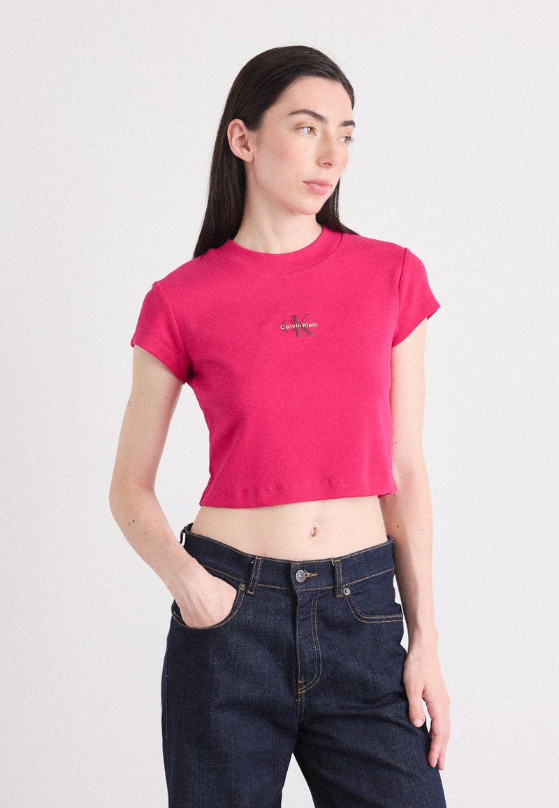 Mladá žena sa pózuje, oblečená v priliehavom ružovom crop topu s logom, skombinovanom s tmavými džínsami. Stojí proti neutrálnej pozadí.