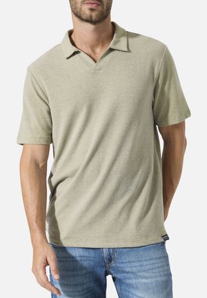 Mann trägt beige kurzärmeliges Poloshirt und blaue Jeans, steht mit einer Hand entspannt und der anderen hinter dem Rücken.