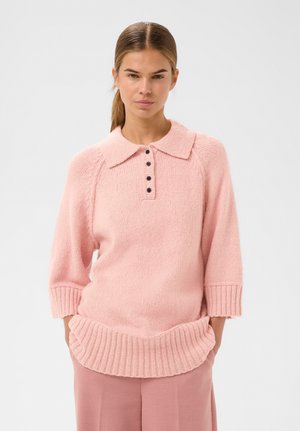 Frau in einem hellrose gestrickten Pullover mit geripptem Saum, Dreiviertelärmel, Kragen und schwarzen Knöpfen, kombiniert mit rosa Hosen.