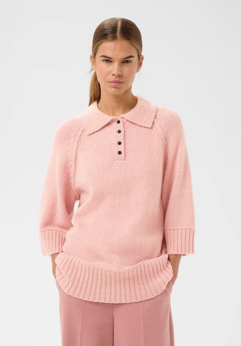 Žena v jasně růžovém pleteném svetru s žebrovaným lemem, tříčtvrtečními rukávy, límečkem a černými knoflíky, v kombinaci s růžovými kalhotami.