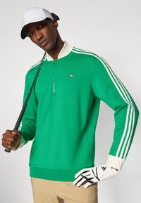 Zelený zipsový dlhý rukáv s bielymi pruhmi, má polo golier a logo. Model drží golfovú palicu a má na sebe biele golfové rukavice.