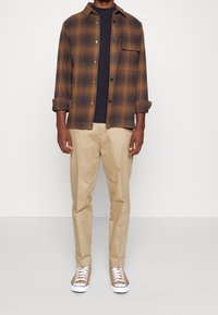 Brun rutig flanellskjorta, svart t-shirt under, beige byxor och bruna sneakers med vita sulor. Knäppt och med uppvikt ärmar.