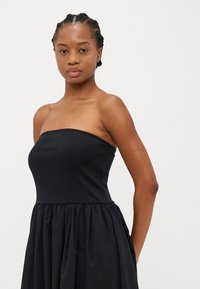 Svart stroppeløs kjole laget av myk bomull, med et tettsittende liv og rynket, flytende skjørt. Minimalistisk design uten dekorasjoner.