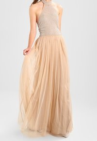 Halter-neck klänning med en texturerad, åtsittande överdel i ljusbeige, som flödar ut i en full, lager-på-lager tullekjol med en mjuk, genomskinlig finish.