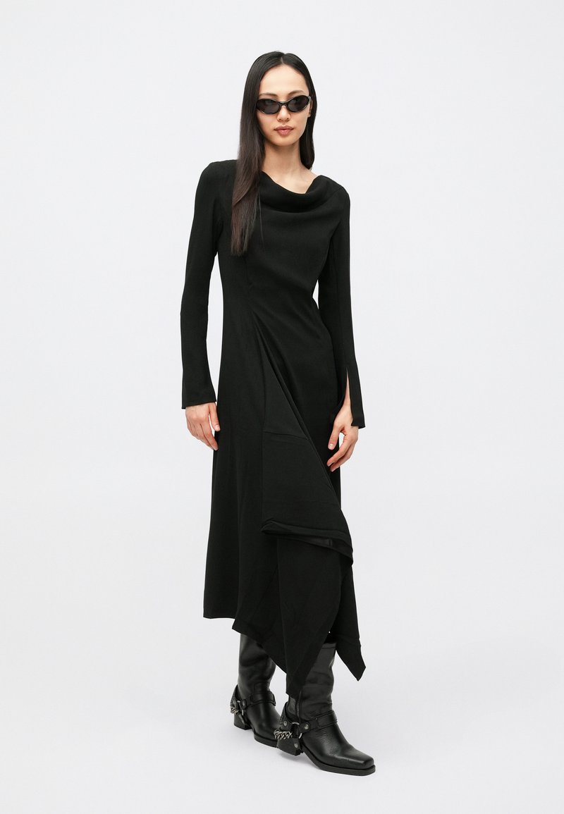 Vestito nero a maniche lunghe con scollo drappeggiato a cappuccio, orlo asimmetrico e spacchi laterali. Abbinato a stivali neri al ginocchio e occhiali da sole.