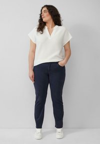 Witte, gestructureerde top met korte mouwen en een V-hals, gecombineerd met navy slim-fit broeken en witte sneakers. Het model staat casual met één hand in een zak.