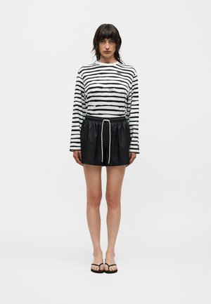 Mujer de cabello oscuro está de pie con una camiseta de manga larga blanca y negra a rayas, pantalones cortos negros con cordón y chanclas negras sobre un fondo blanco.