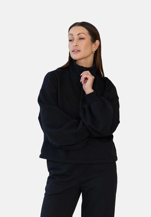 Kobieta z długimi brązowymi włosami, ubrana w czarną bluzę na zamek i spodnie, pozująca z jedną ręką przy brodzie, patrząca na bok.