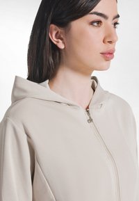 Sudadera con capucha beige con cremallera, de textura suave y ajuste holgado. Cuenta con capucha con cordón y un detalle de cremallera plateada en el frente.
