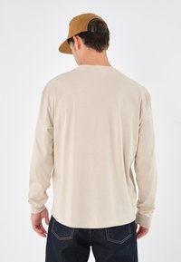 Beige skjorte med lange ermer laget av myk stoff, med rund hals og avslappet passform, kombinert med mørk denimbukse og en brun caps.